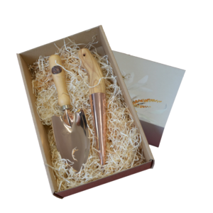 Geschenkbox mit einer Kupferkelle und einem Pflanzenstecher, umgeben von Holzspänen, in einer dekorativen Verpackung.